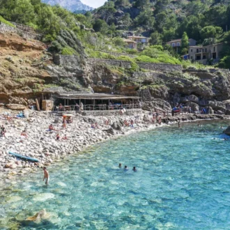Pebble beach and turquoise water at Cala Deià beneath pine-clad cliffs in Mallorca’s Tramuntana.